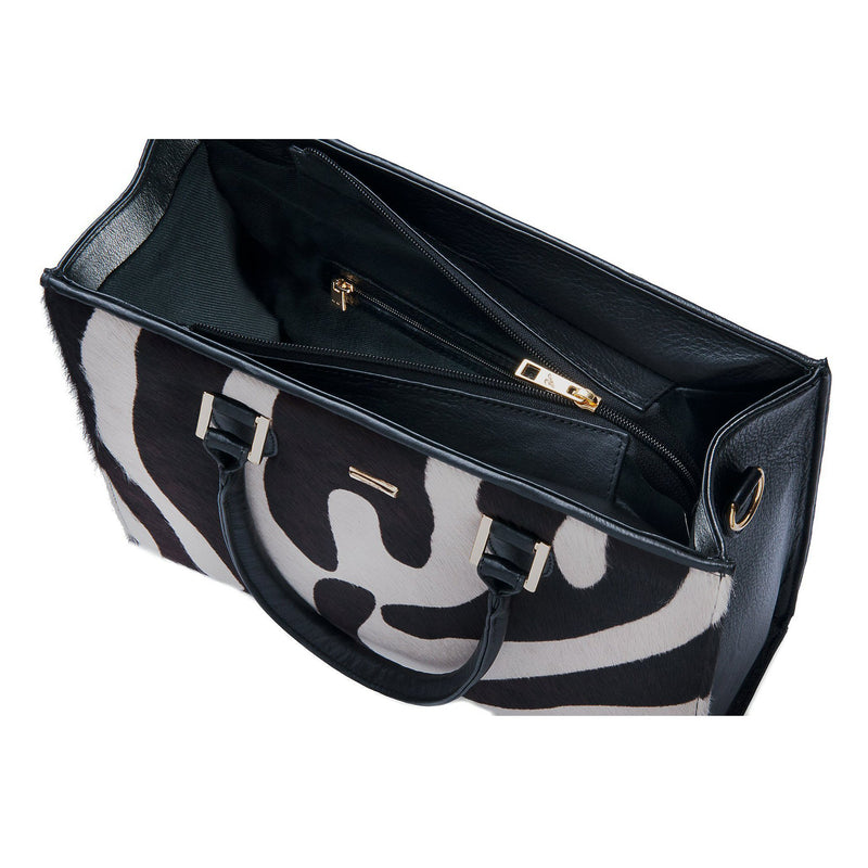 Linda side front top view. Black bovine leather side, back and bottom panel, with white and black zebra printed hair on hide on the front panel. Contains an inside zipper with a back-zip pocket. The bag interior consists of high-quality black cotton suede lining. Includes a top zip closure with gold plated hardware, and a detachable shoulder strap with an adjustable buckle. Bag feet studs are located on the bottom panel.