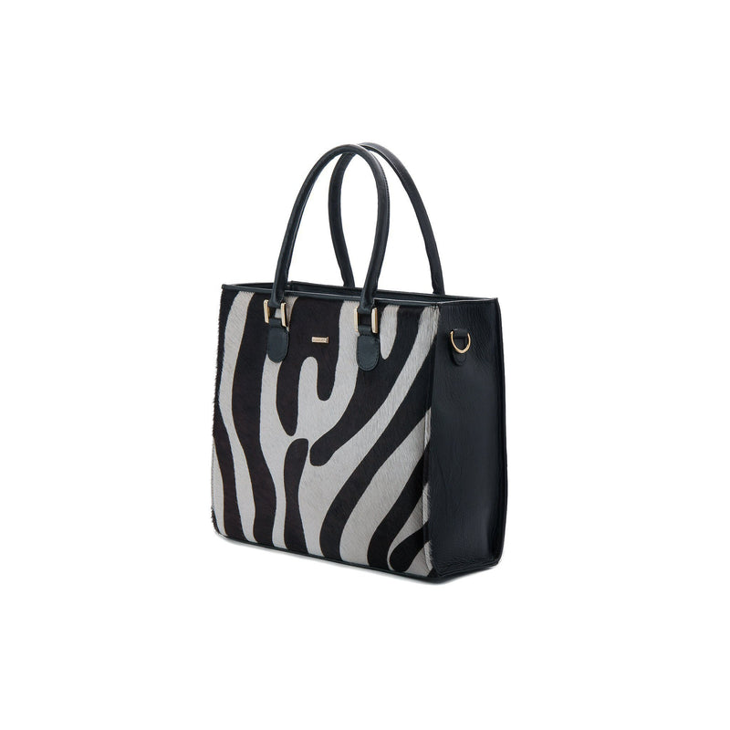 Linda side front view. Black bovine leather side, back and bottom panel, with white and black zebra printed hair on hide on the front panel. Contains an inside zipper with a back-zip pocket. The bag interior consists of high-quality black cotton suede lining. Includes a top zip closure with gold plated hardware, and a detachable shoulder strap with an adjustable buckle. Bag feet studs are located on the bottom panel.